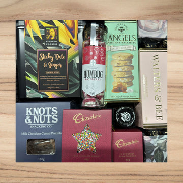 Assorted gourmet snacks including Sticky Date & Ginger cookies, raspberry humbug, salted caramel nougat biscuits, chocolate-coated pretzels, and Christmas plum puddings on wood