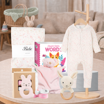 Baby gift set arranged on a round table in a softly styled nursery or living room. The set includes a cream one-piece baby outfit on a wooden hanger, a pink plush bunny lovey, a white and pink patterned blanket, a boxed baby teether, a small plush toy, a book titled "Bebe's First Words," and a knitted bunny teether with a wooden ring.