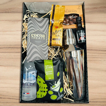 Gift basket with drinks, snacks, and a cocktail shaker on a wooden surface