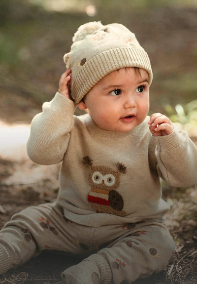 Baby wearing a cream knit hat and sweater with a reindeer design, sitting outdoors.
