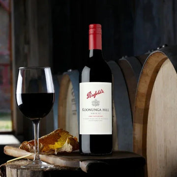 Penfolds Koonunga Hill wine bottle with a glass of red wine and bread on a wooden barrel.