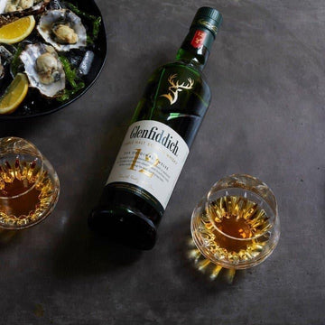 Glenfiddich whisky bottle with two glasses and oysters on a dark surface