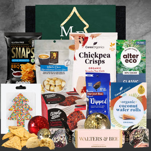 Assorted gluten-free snacks and treats including Snaps BBQ chips, White Choc Macadamia Cookie Bites, organic chickpea crisps, dark chocolate, and Christmas pudding