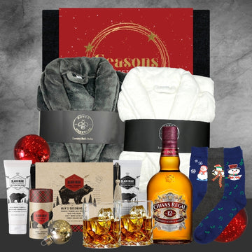 Holiday gift set with gray and white luxury bathrobes, Chivas Regal whisky, Black Bear grooming products, festive socks, and Christmas ornaments