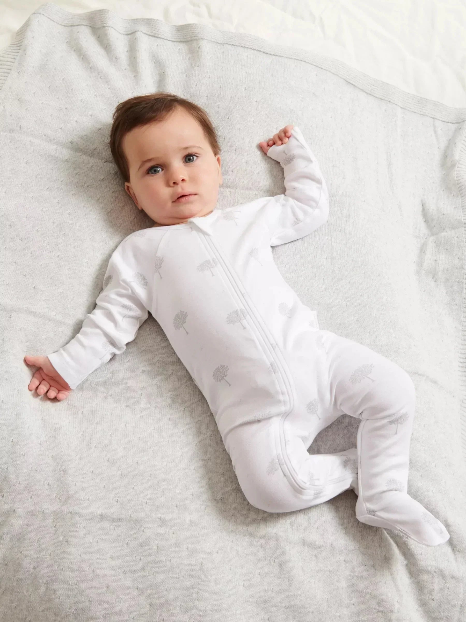 Baby lying on a gray blanket wearing a white onesie with small patterns.