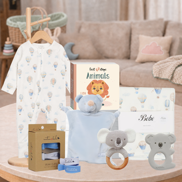 A baby gift set arranged on a round wooden table in a softly styled nursery setting. The set includes a white long-sleeve baby jumpsuit with a pastel hot air balloon print hanging on a wooden hanger, a book titled 'Animals', a light blue plush bear security blanket, a koala teether ring, a koala pacifier clip, a box of baby wipes, and a boxed item labeled 'Bebe'.