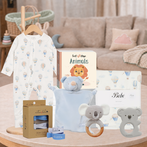 A baby gift set arranged on a round wooden table in a softly styled nursery setting. The set includes a white long-sleeve baby jumpsuit with a pastel hot air balloon print hanging on a wooden hanger, a book titled 'Animals', a light blue plush bear security blanket, a koala teether ring, a koala pacifier clip, a box of baby wipes, and a boxed item labeled 'Bebe'.