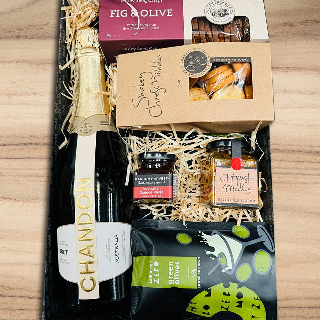 Gift basket with Chandon champagne, assorted snacks, and fig and olive products on a wooden surface.