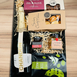 Gift basket with Chandon champagne, assorted snacks, and fig and olive products on a wooden surface.