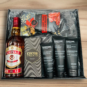 Gift set with Chivas Regal whiskey, chocolate bars, and grooming products on a wooden surface.