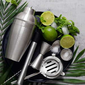 Cocktail shaker and tools with limes, mint, and ice on a dark surface with green leaves.