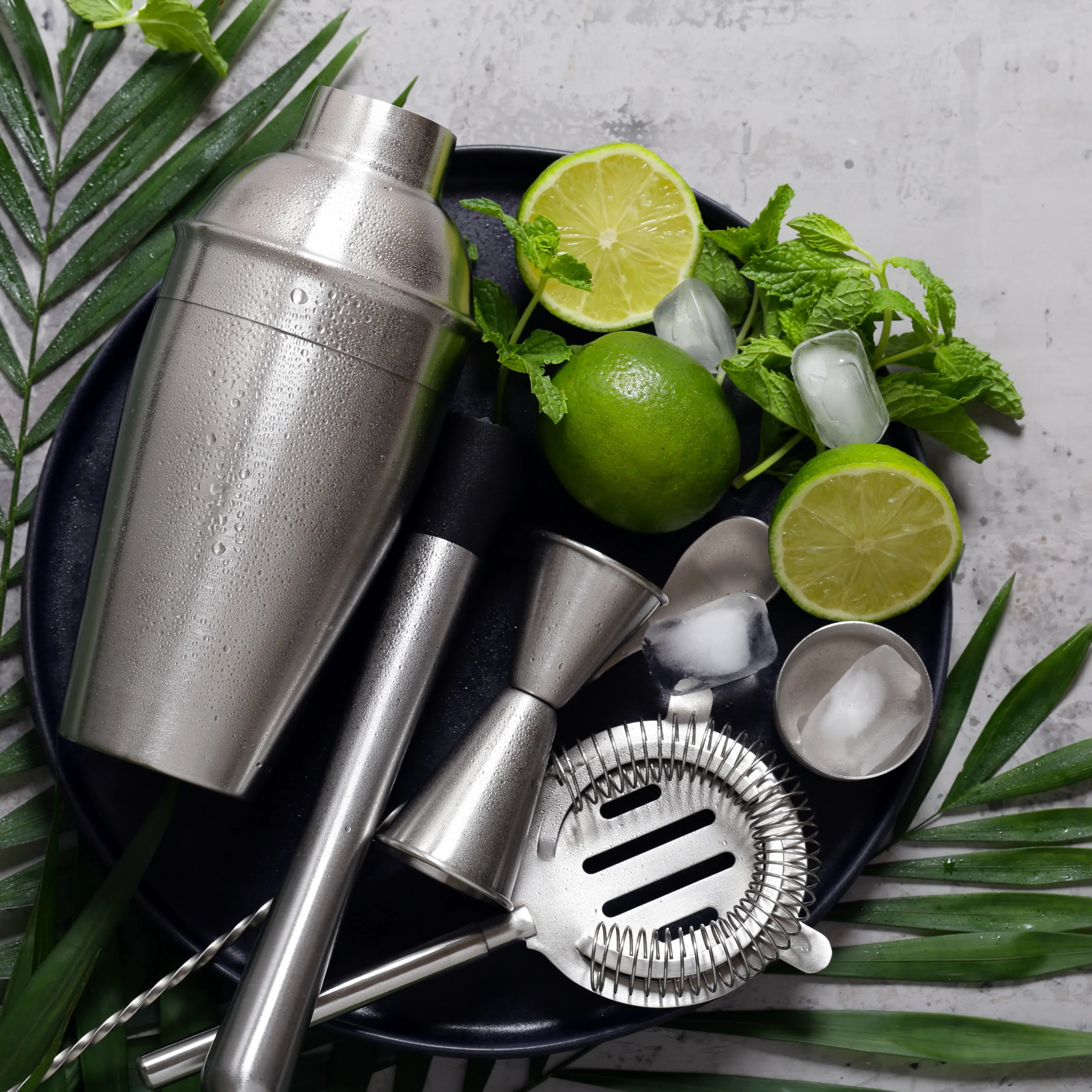 Cocktail shaker and tools with limes, mint, and ice on a dark surface with green leaves.