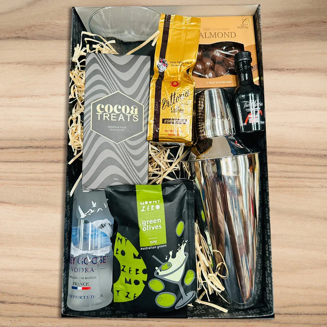 Gift basket with drinks, snacks, and a cocktail shaker on a wooden surface