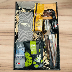 Gift basket with drinks, snacks, and a cocktail shaker on a wooden surface