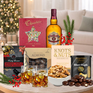 Gourmet gift set with Chivas Regal whiskey, chocolates, and snacks on a table with a festive background.