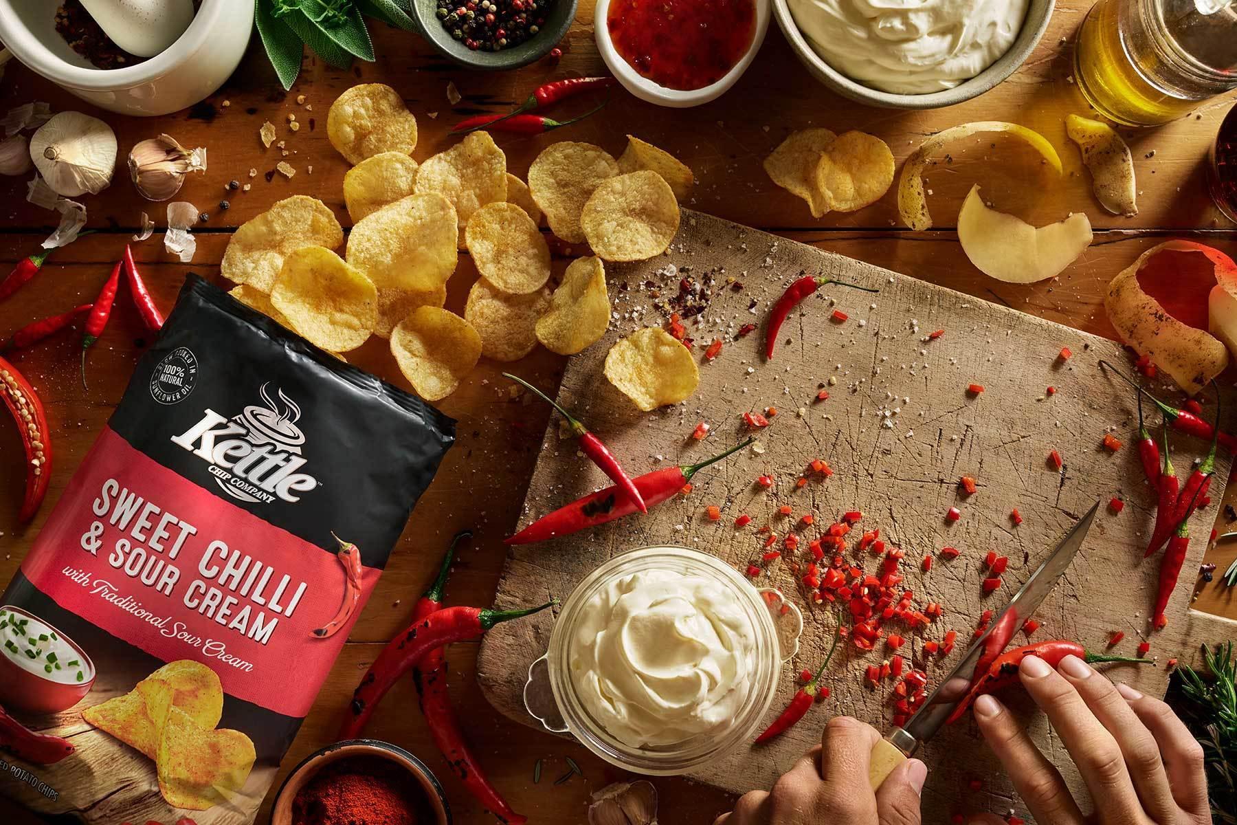 Kettle Sweet Chilli & Sour Cream potato chips on a wooden board with ingredients and a person preparing food.
