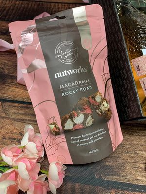 Pink and brown packaging of Nutworks Macadamia Rocky Road on a wooden surface with flowers.