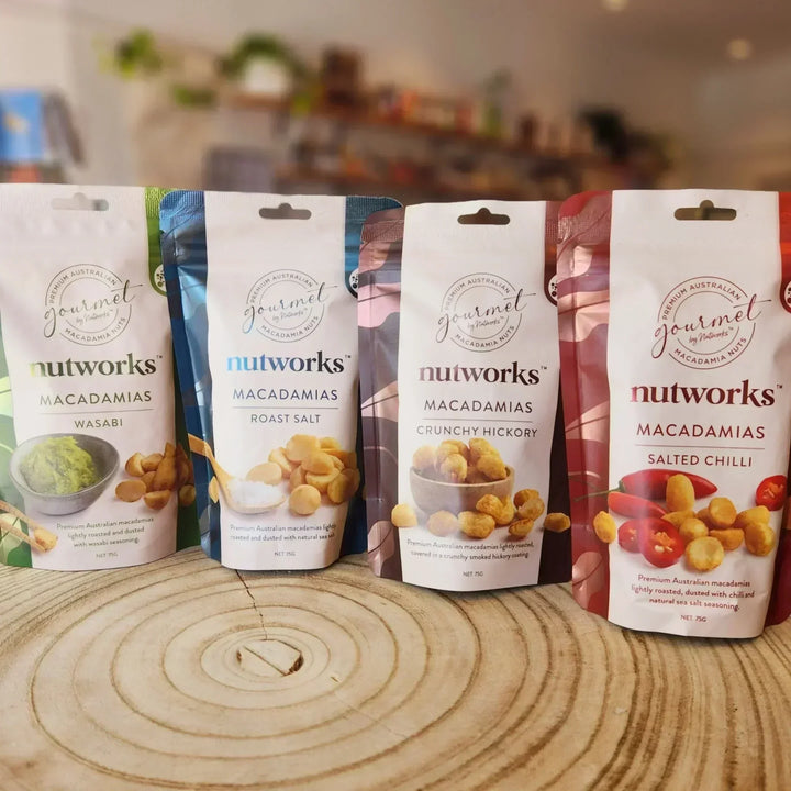 Five packages of Nutworks macadamias on a wooden surface with a blurred kitchen background.