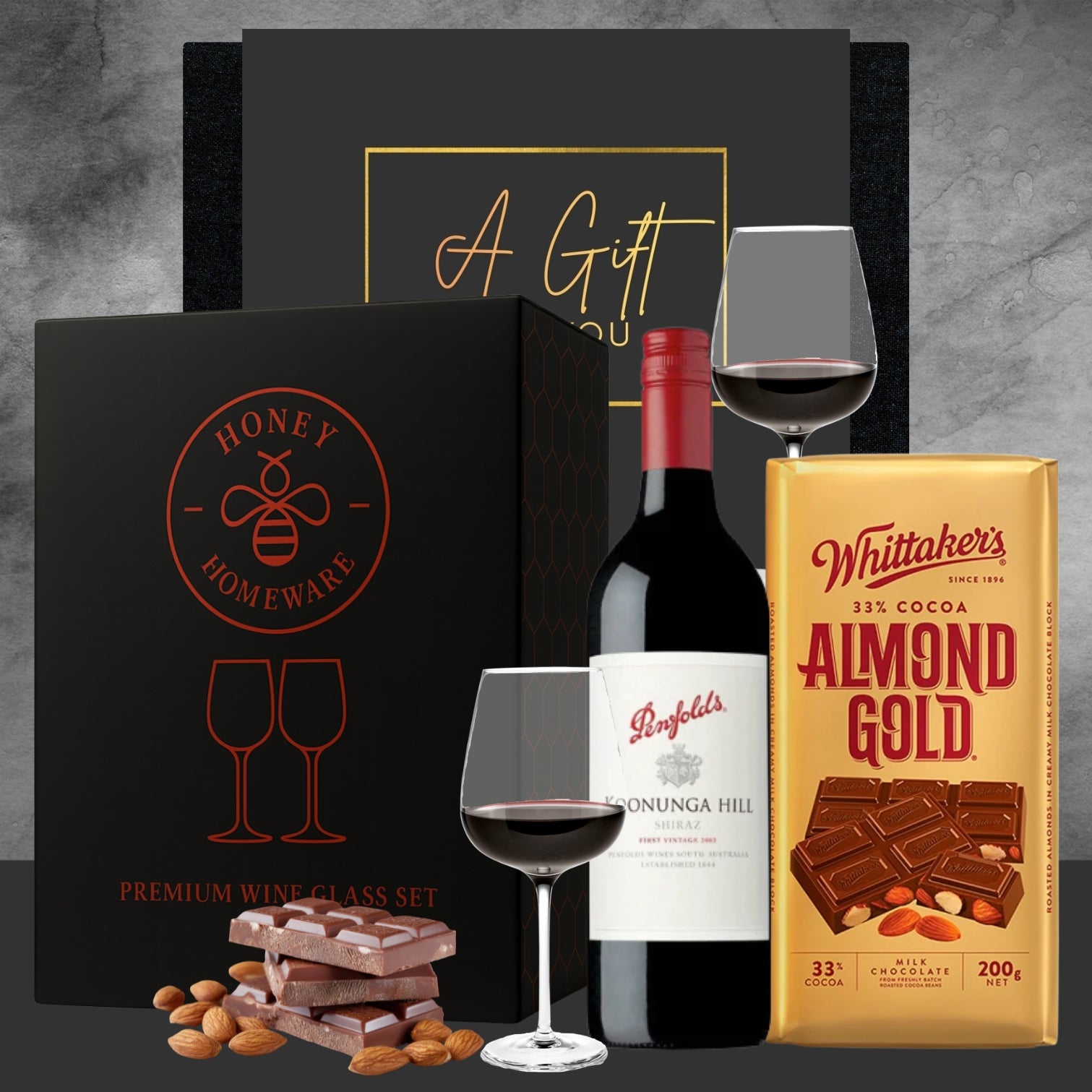 Penfolds wine bottle with a glass, Honey & Homeware box, and Whittaker's Almond Gold chocolate on a gray background.