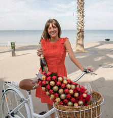 Woman in a red dress holding a bottle of wine with a bicycle and basket of fruit on a beach.