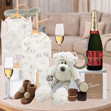 A baby gift hamper set arranged on a round table in a softly blurred living room. The set includes a striped and printed baby onesie, a plush white teddy bear, brown knit baby booties, two champagne flutes, a bottle of Krug champagne, and chocolate treats.