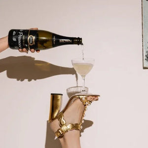 Bottle of champagne being poured into a glass with a hand holding a high-heeled shoe.