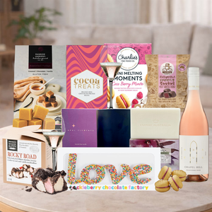 Assorted gift items including chocolate, wine, and snacks on a table with a blurred background.