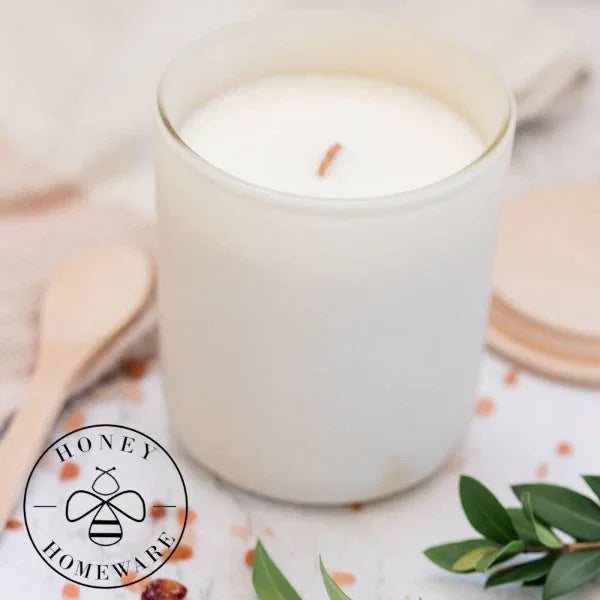 White candle in a glass jar with a wooden lid on a decorative surface, featuring the Honey Homeware logo.