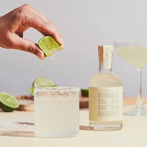 Person squeezing lime into a glass of margarita with a bottle of tequila in the background.