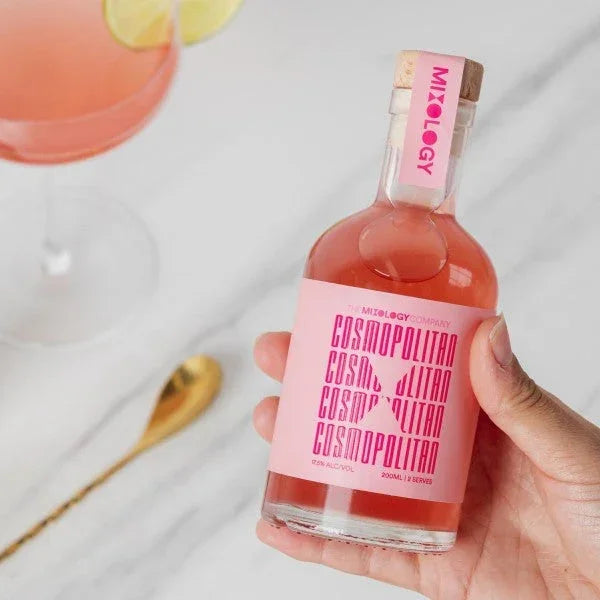 Bottle of pink cocktail mixer with 'Mixology' branding held by a hand, with a glass of pink drink and lemon slices in the background.