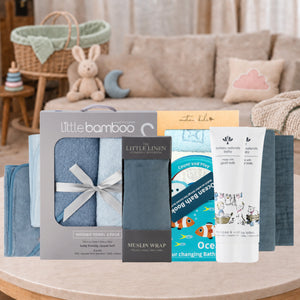 A baby gift set displayed on a round wooden table in a cozy nursery setting. The set includes a boxed hooded towel pack by Little Bamboo, a muslin wrap, a bath book, and a bath sponge, all arranged in front of a soft-focus living room background with a bunny toy and cushions.