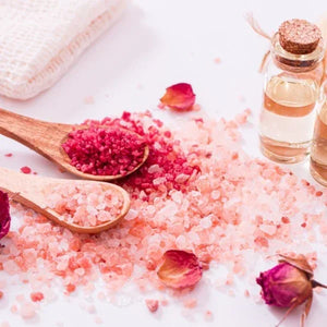 Pink bath salts with wooden spoons and rose petals on a white surface