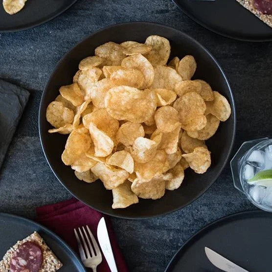 Black bowl filled with potato chips on a dark surface with plates and cutlery around.