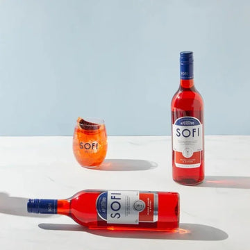 Two bottles and a glass of SOFI aperitivo on a white surface with a light blue background