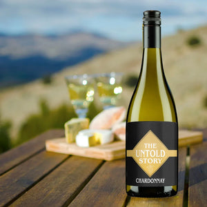 Bottle of Chardonnay with 'The Untold Story' label on a wooden table with a scenic background.