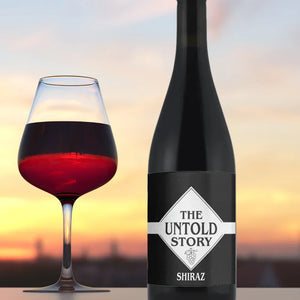 Wine bottle and glass of red wine with 'The Untold Story Shiraz' label against a sunset background.