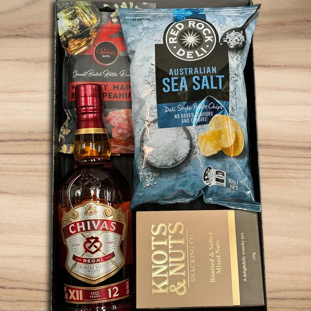 Chivas Regal bottle, Red Rock Deli Australian Sea Salt chips, and Knott's Nuts snack mix in a box on a wooden surface.