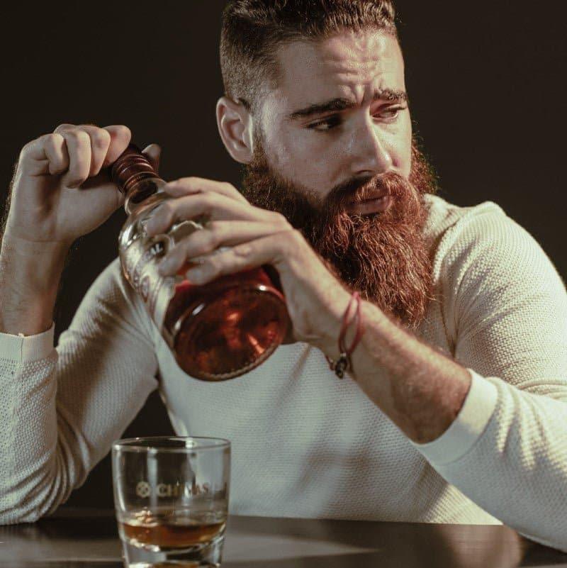 Man with a beard pouring whiskey into a glass against a dark background