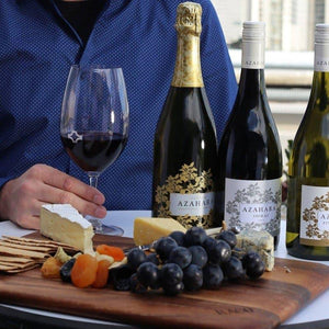 Wine bottles and a glass of red wine on a table with a person in a blue shirt, surrounded by snacks.