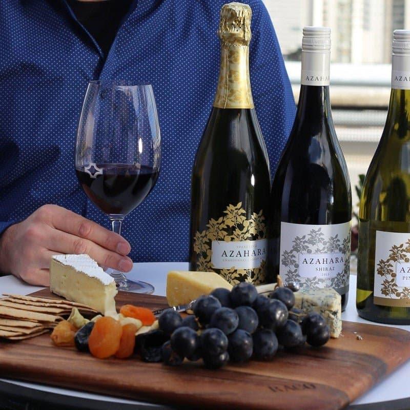 Wine bottles and a glass of red wine on a table with a person in a blue shirt, surrounded by snacks.