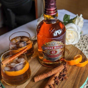 Bottle of Chivas Regal 12 blended Scotch whisky with two glasses, cinnamon sticks, star anise, and orange peel on wooden board