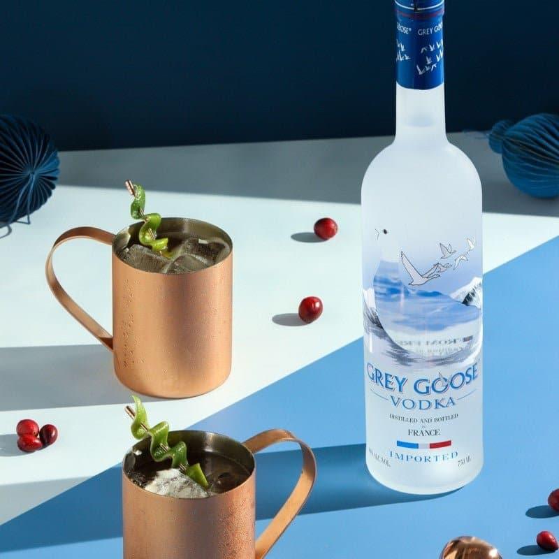 Bottle of Grey Goose vodka with two copper mugs on a blue and white geometric background