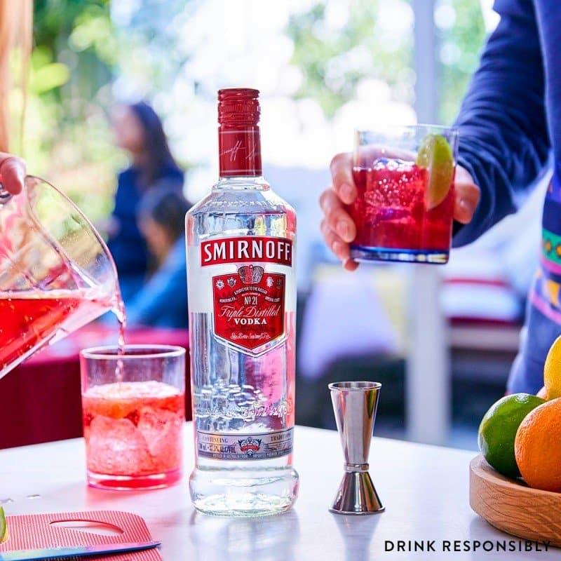 Smirnoff vodka bottle with a cocktail being poured and held, set against a blurred social setting.